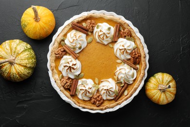 Delicious homemade pumpkin pie in baking dish and fresh pumpkins on black textured table, flat lay