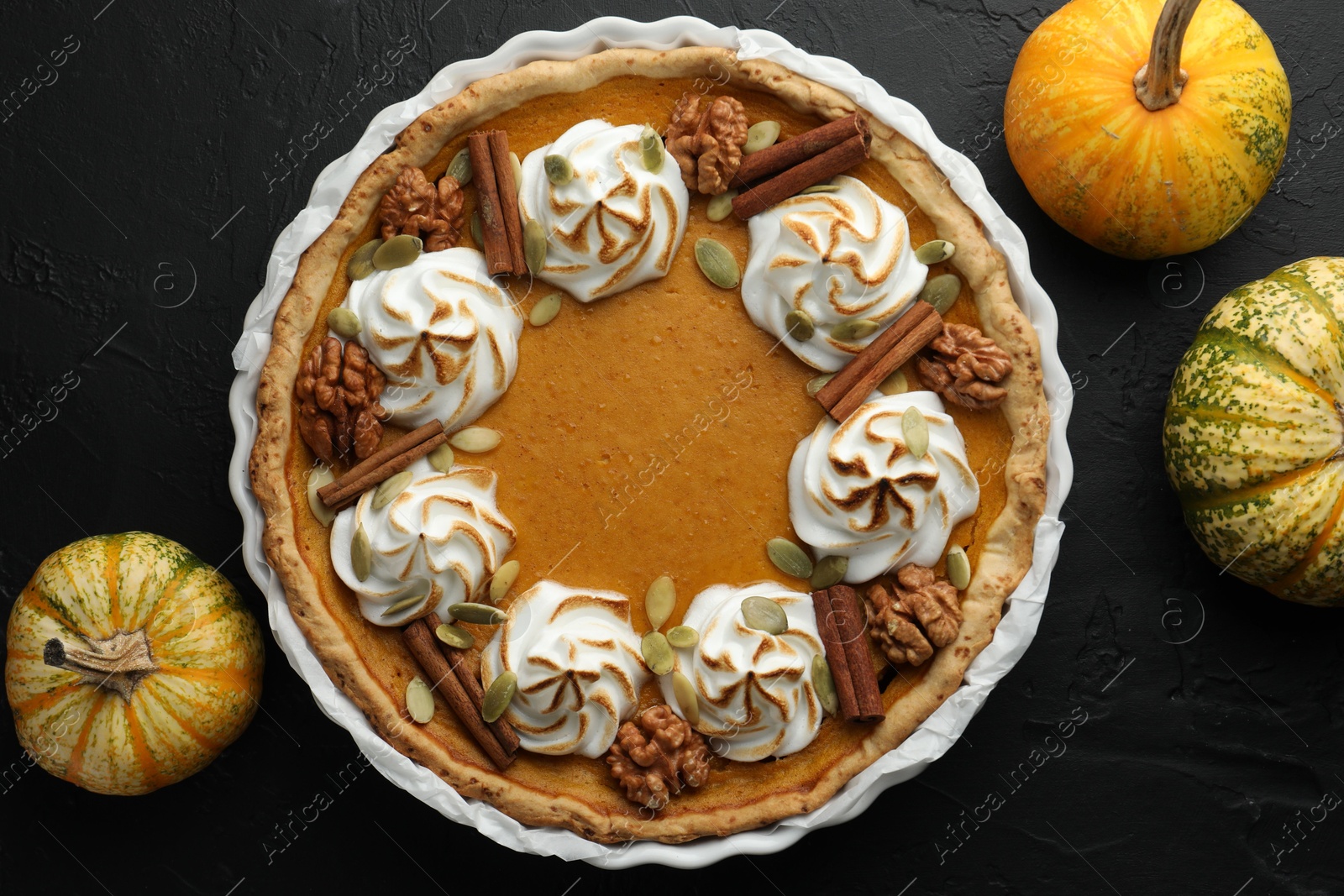 Photo of Delicious homemade pumpkin pie in baking dish and fresh pumpkins on black textured table, flat lay