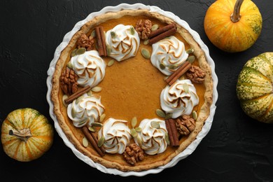 Photo of Delicious homemade pumpkin pie in baking dish and fresh pumpkins on black textured table, flat lay