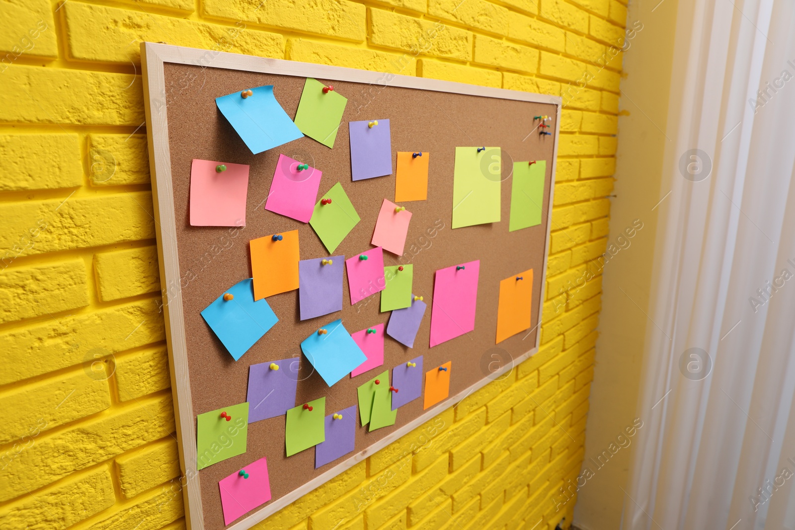 Photo of Corkboard filled with colorful notes and pins on yellow brick wall indoors