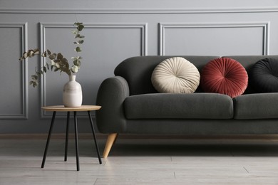 Photo of Stylish sofa with cushions and eucalyptus branches in vase on coffee table near grey wall