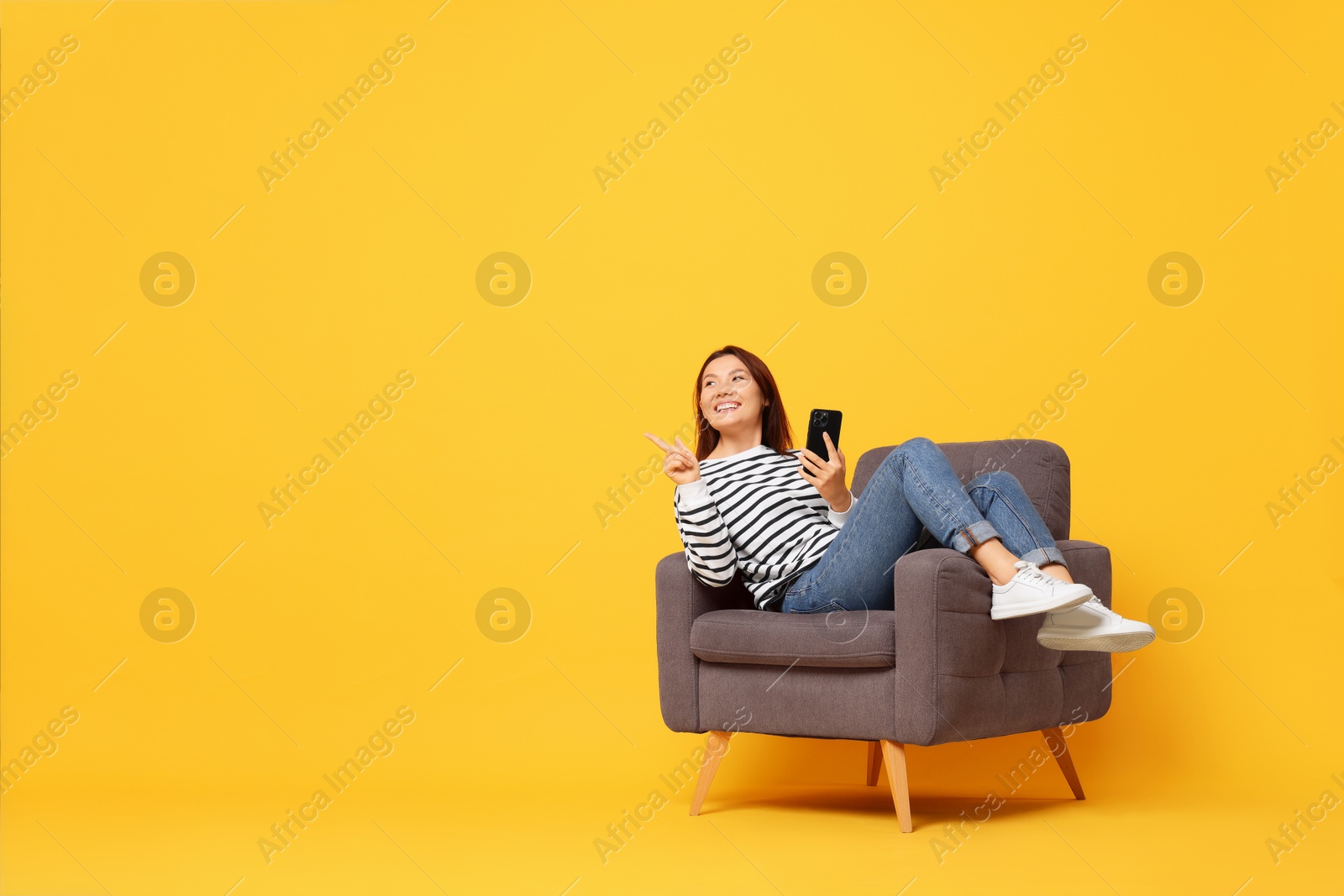 Photo of Smiling woman with smartphone sitting in armchair on yellow background. Space for text