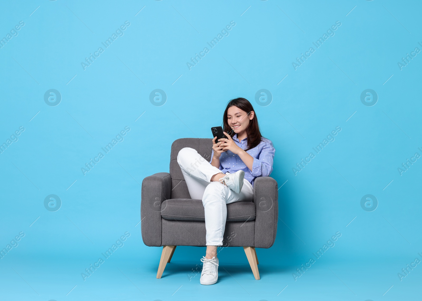 Photo of Smiling woman with smartphone resting in armchair on light blue background