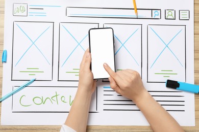 Photo of Website designer using smartphone at wooden table with wireframe and stationery, top view