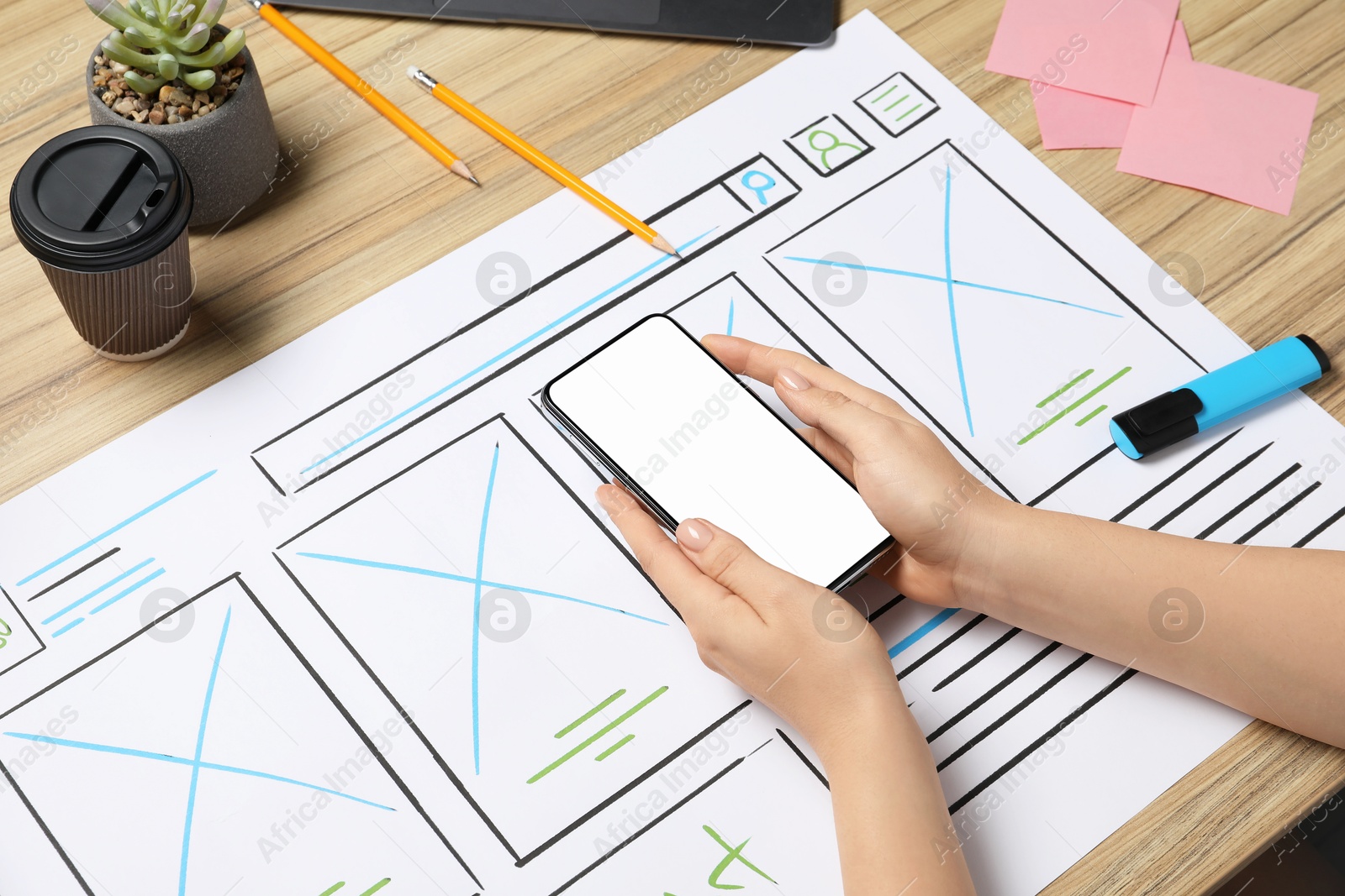 Photo of Website designer holding smartphone at wooden table with wireframe and stationery, closeup