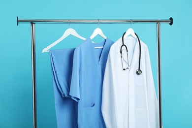 Photo of Different medical workers' uniforms and stethoscope on clothing rack against light blue background