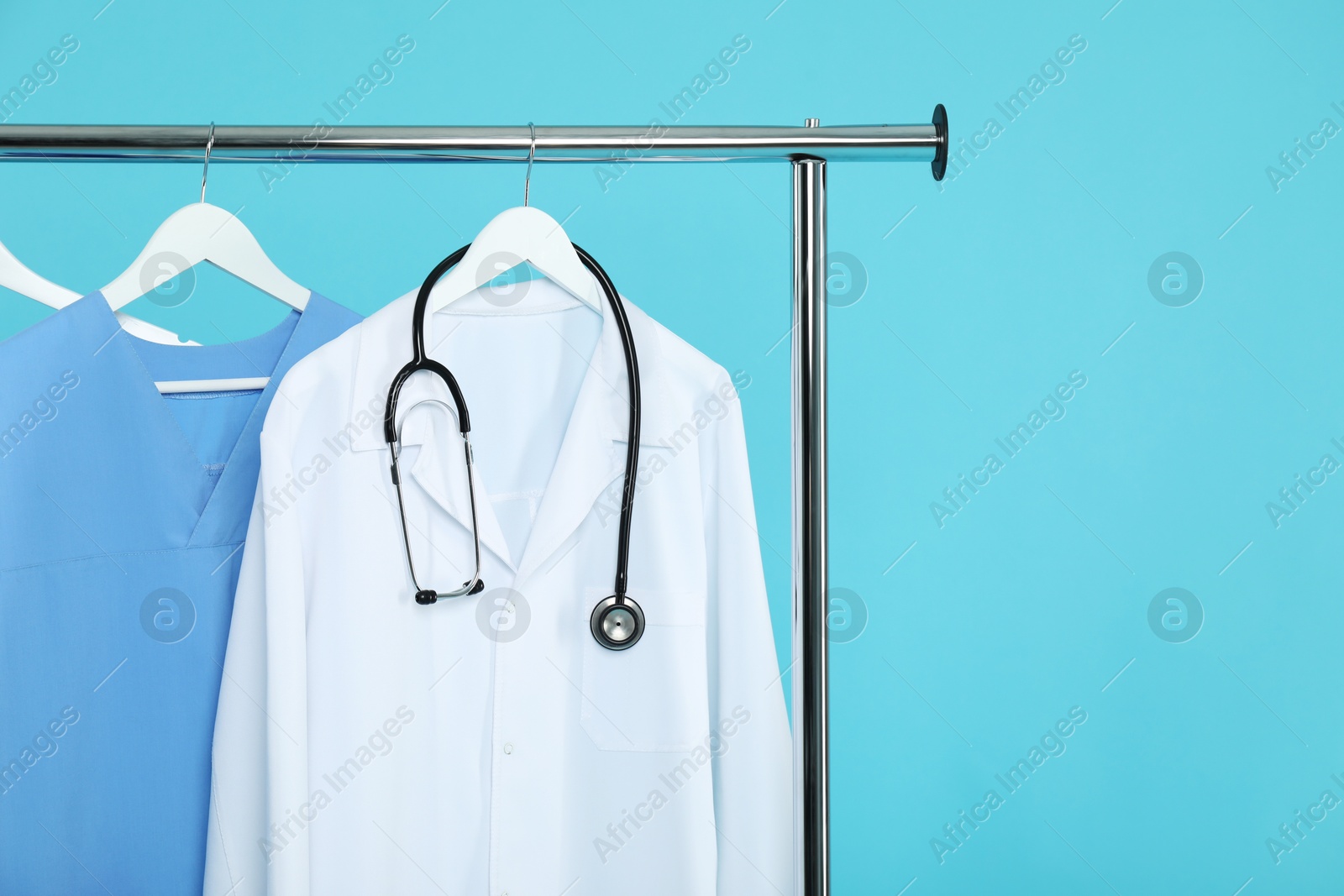 Photo of Different medical workers' uniforms and stethoscope on clothing rack against light blue background, space for text