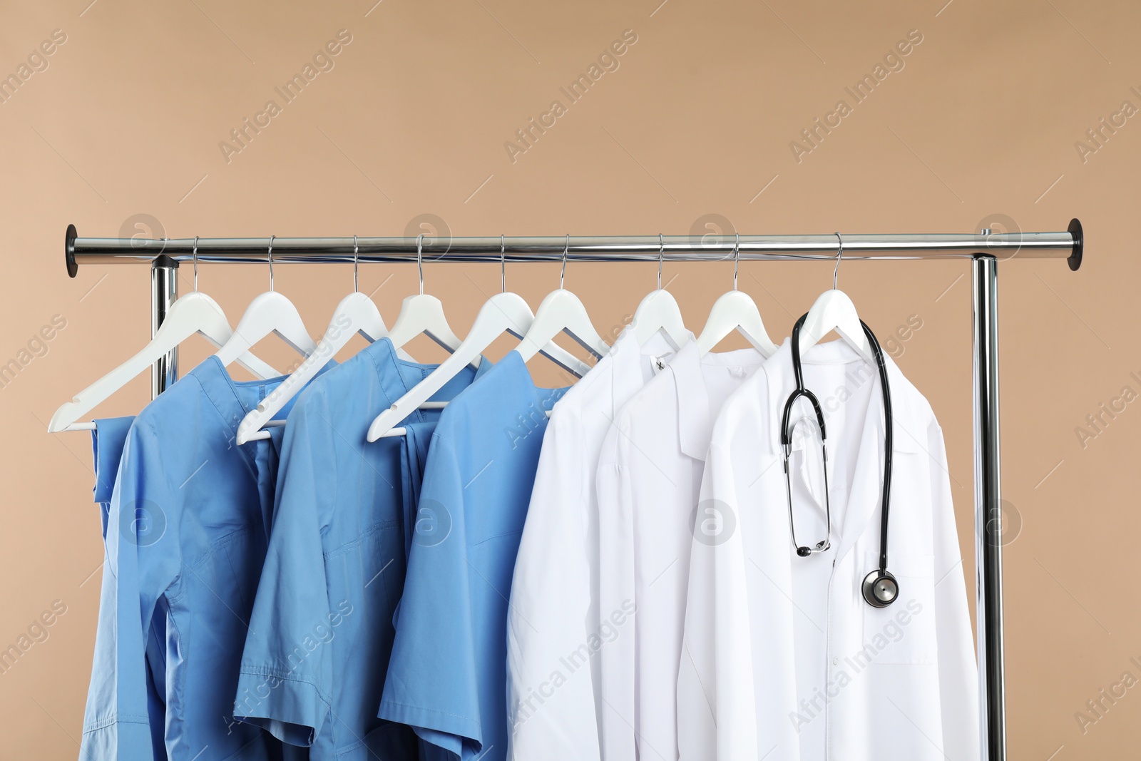 Photo of Different medical workers' uniforms and stethoscope on clothing rack against beige background