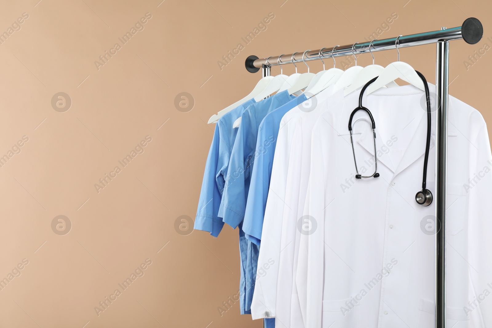 Photo of Different medical workers' uniforms and stethoscope on clothing rack against beige background, space for text