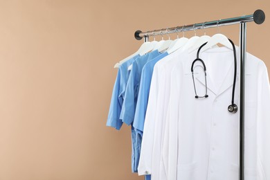 Photo of Different medical workers' uniforms and stethoscope on clothing rack against beige background, space for text