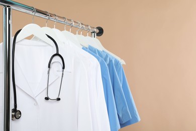 Photo of Different medical workers' uniforms and stethoscope on clothing rack against beige background, space for text