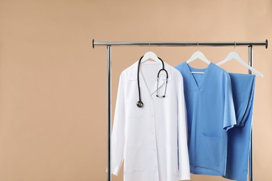 Different medical workers' uniforms and stethoscope on clothing rack against beige background, space for text