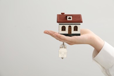 Photo of Woman holding key with keychain and house model on light grey background, closeup. Space for text