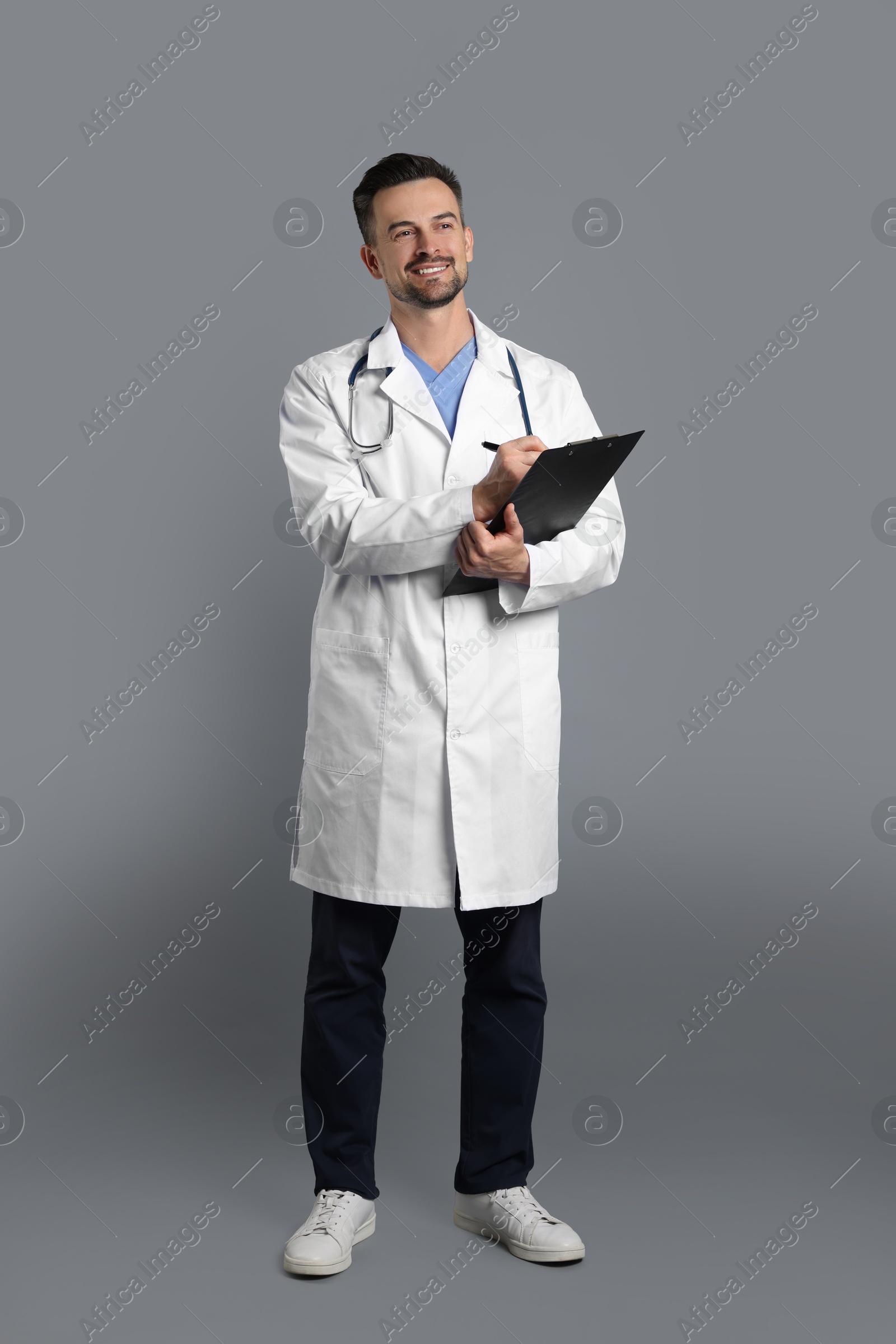 Photo of Smiling doctor with stethoscope and clipboard on grey background