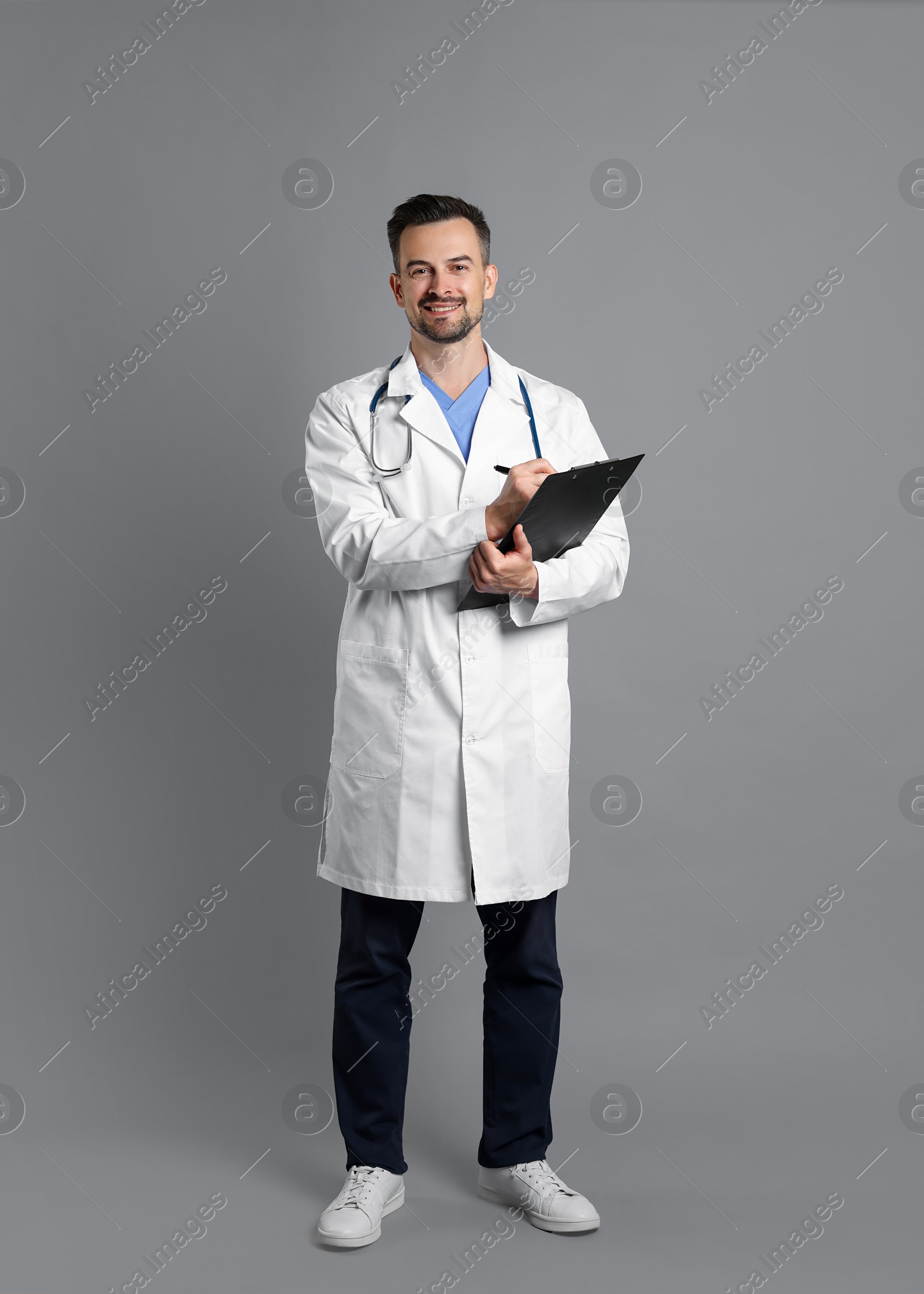 Photo of Smiling doctor with stethoscope and clipboard on grey background