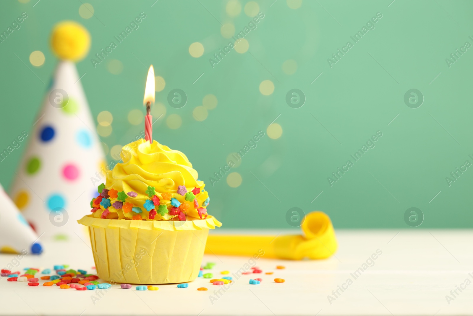 Photo of Tasty cupcake with burning candle and party cones on green background with blurred lights, space for text