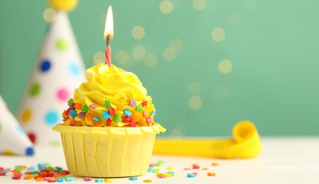 Photo of Tasty cupcake with burning candle and party cones on green background with blurred lights, space for text