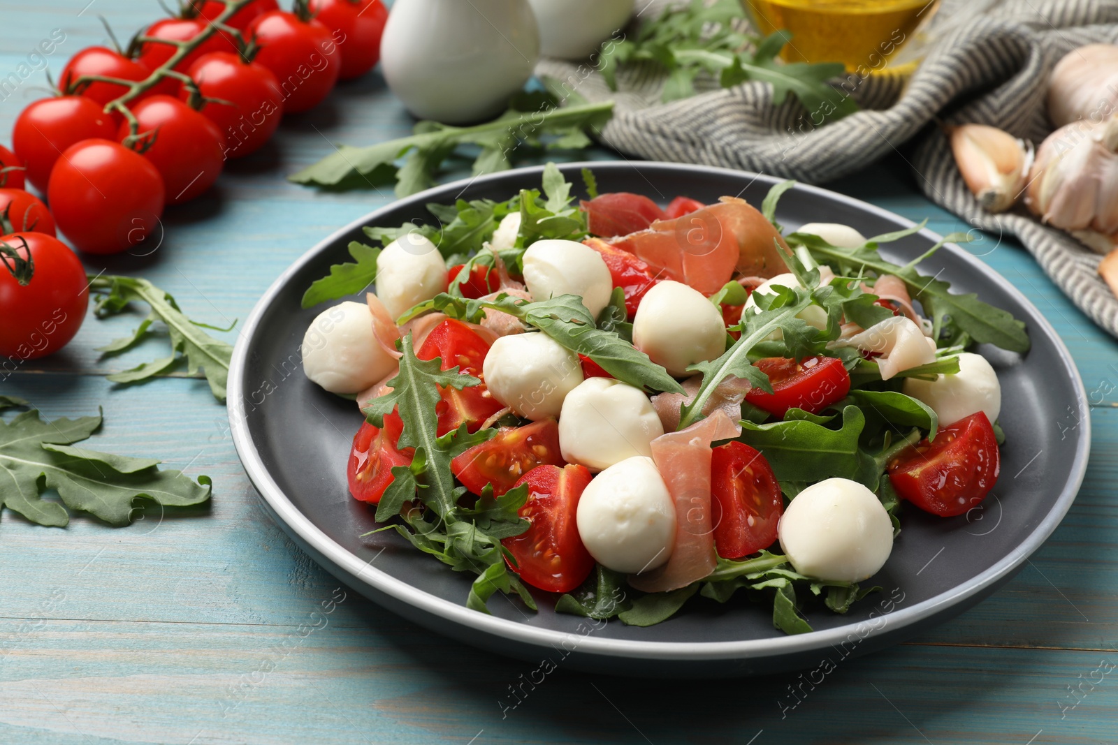 Photo of Tasty salad with arugula, mozzarella cheese, tomatoes and jamon on light blue wooden table, closeup