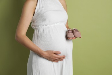 Photo of Young pregnant woman with pair of baby shoes on light olive background, closeup