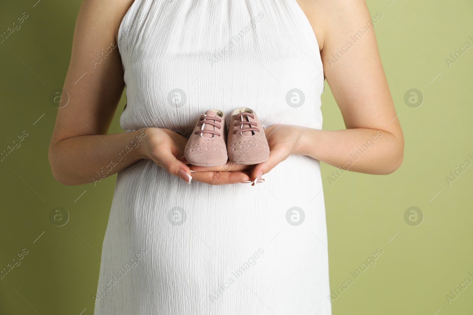 Photo of Young pregnant woman with pair of baby shoes on light olive background, closeup