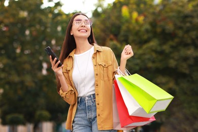 Photo of Happy woman with colorful shopping bags using smartphone outdoors