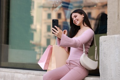 Photo of Happy woman with colorful shopping bags taking selfie outdoors, space for text