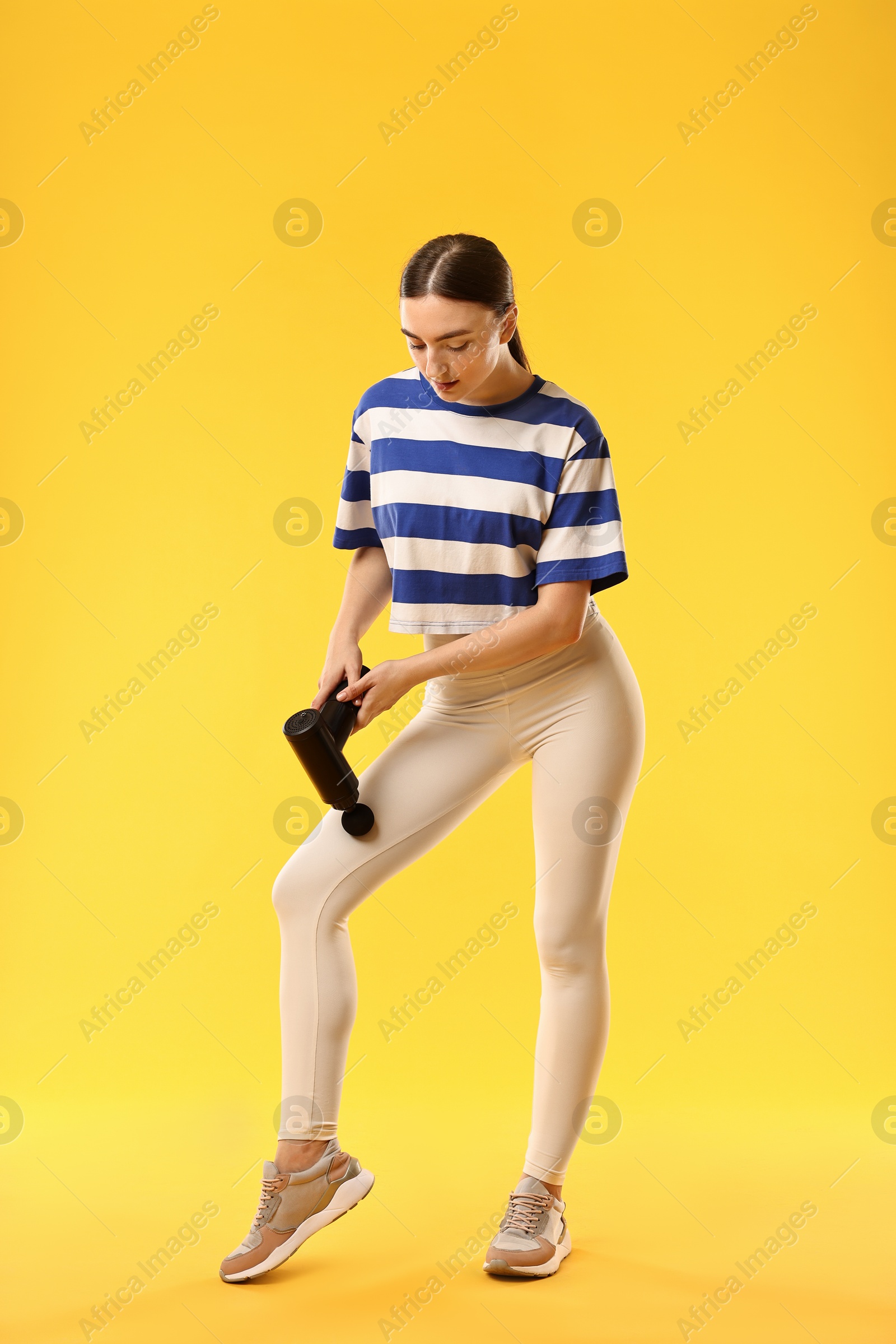 Photo of Woman using percussive massager to relax thigh muscles on yellow background