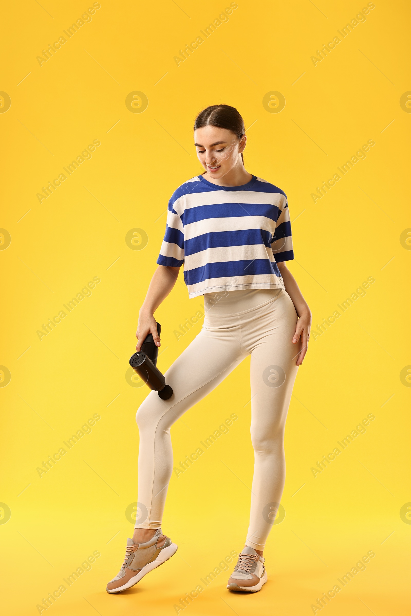 Photo of Woman using percussive massager to relax thigh muscles on yellow background