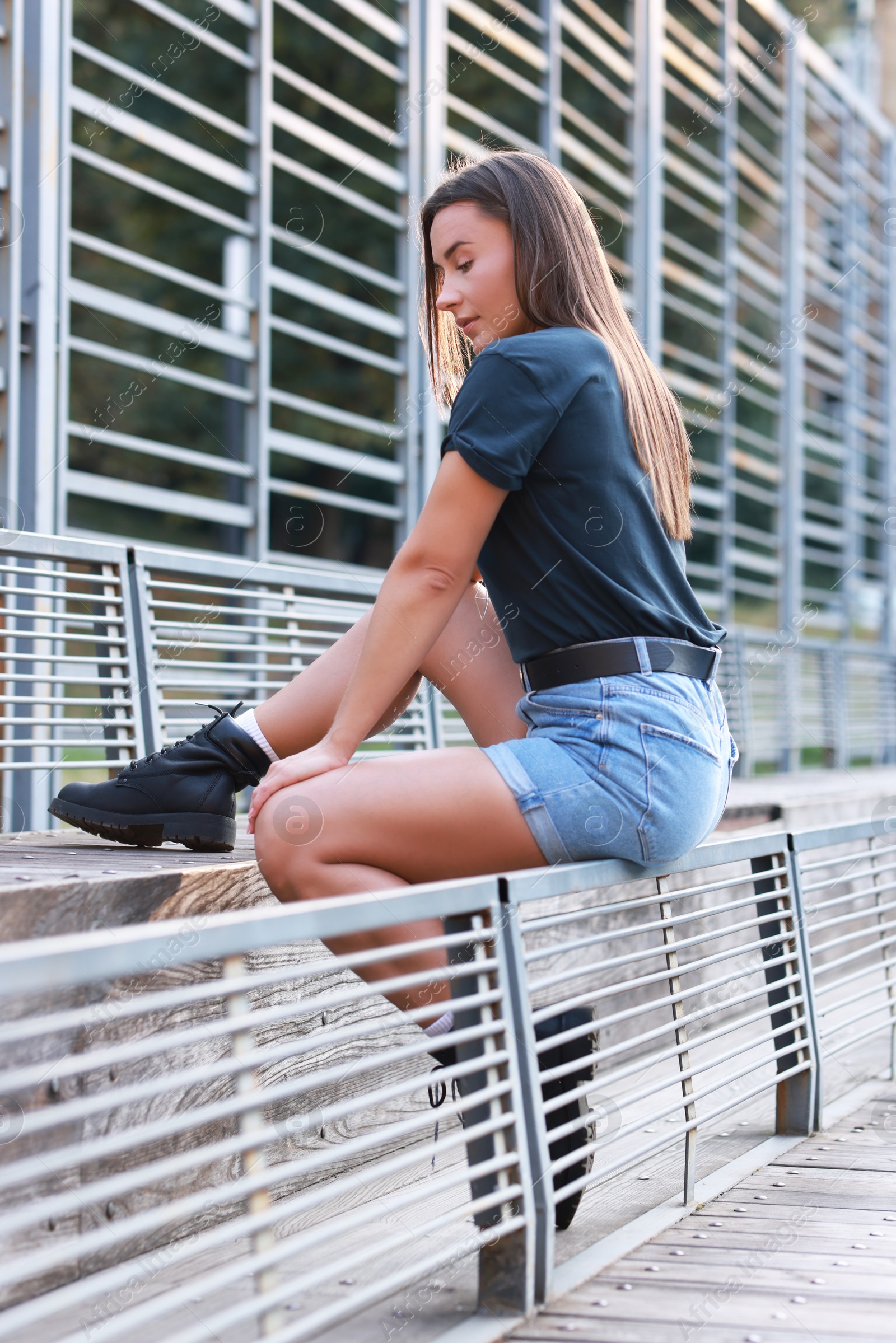 Photo of Beautiful woman in stylish denim shorts sitting on metal bench backrest outdoors