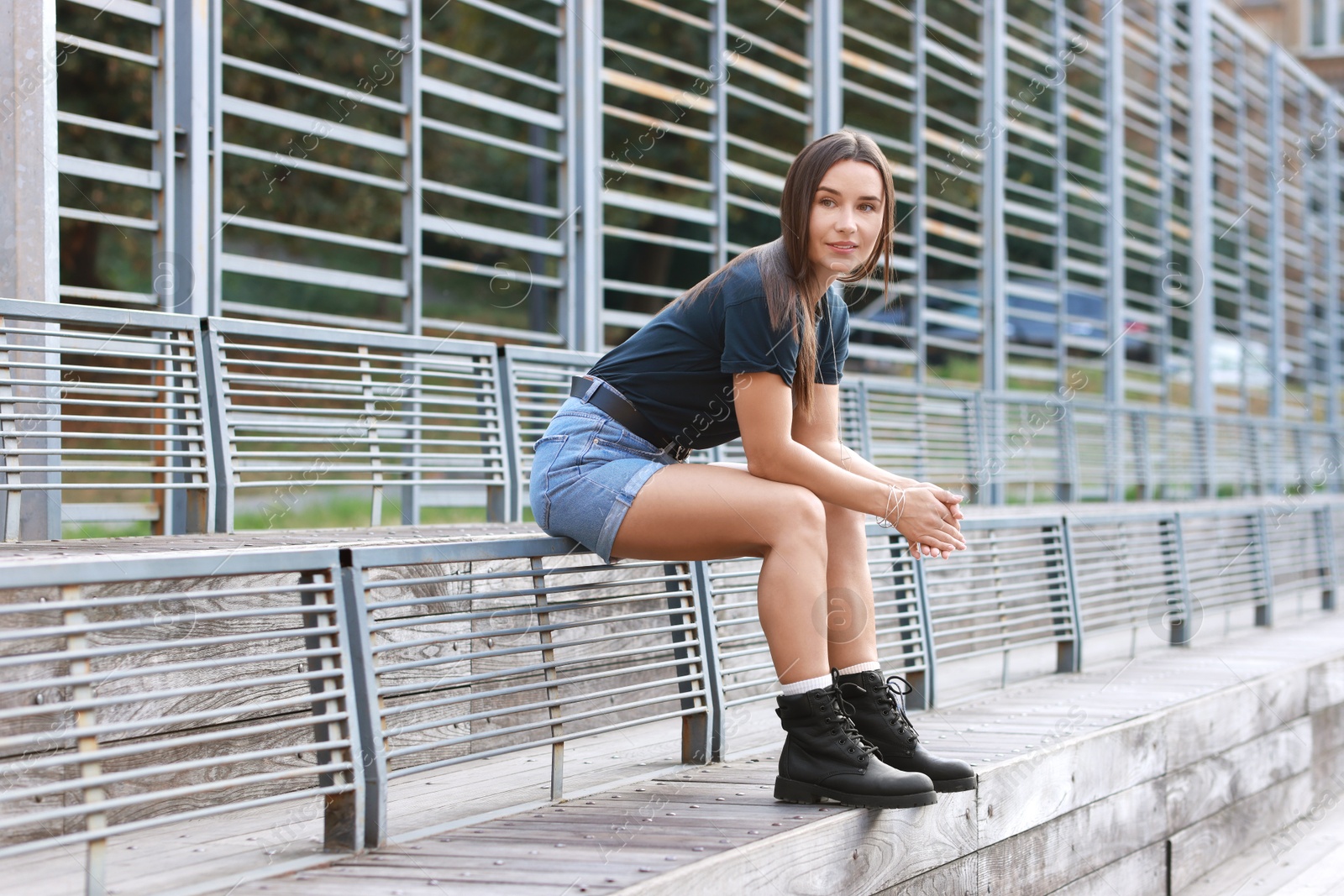 Photo of Beautiful woman in stylish denim shorts sitting on metal bench backrest outdoors