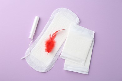 Menstrual pad with red feather and tampon on violet background, top view