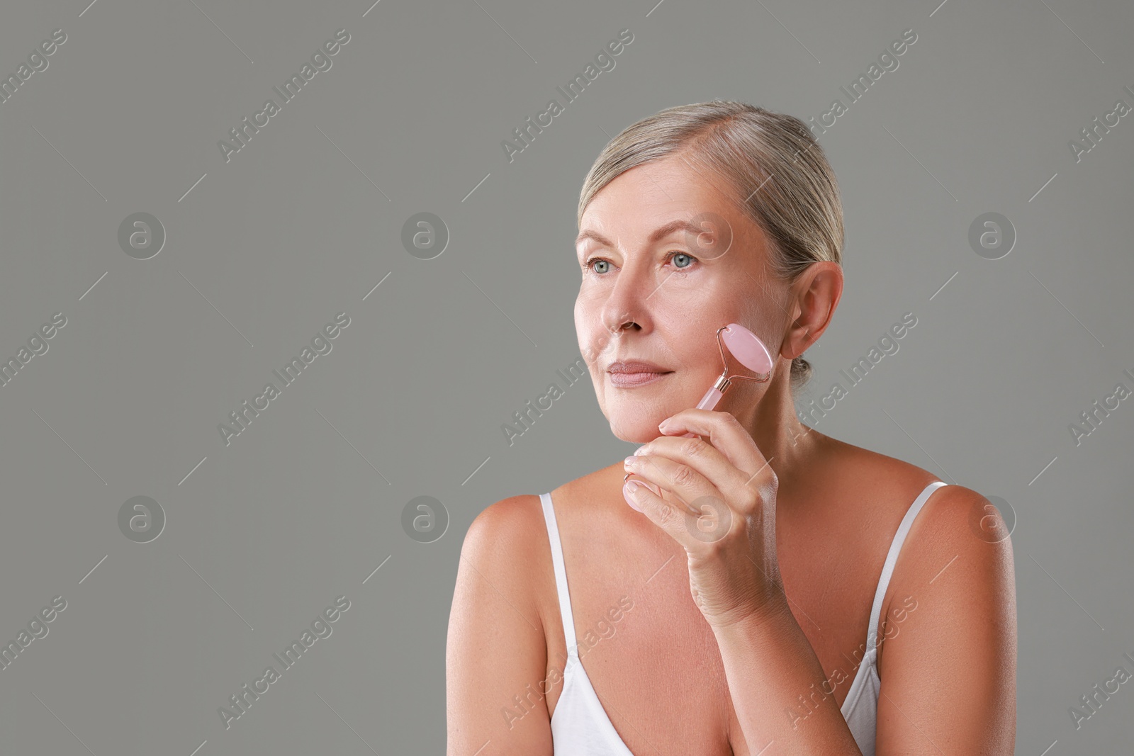 Photo of Beautiful woman doing facial massage with roller on grey background, space for text