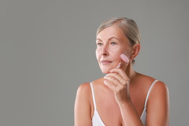 Beautiful woman doing facial massage with roller on grey background, space for text