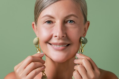 Photo of Beautiful woman doing facial massage with rollers on light green background, closeup