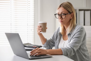 Budget planning. Woman with paper cup of coffee using laptop at table indoors