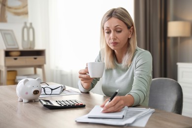 Budget planning. Woman with cup of drink taking notes at table indoors