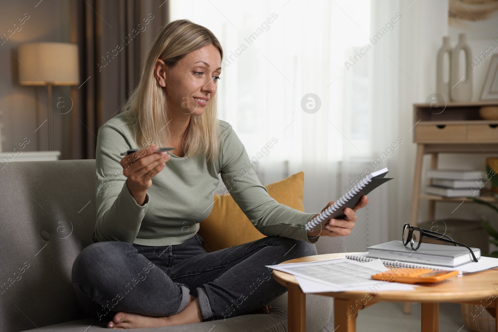 Photo of Budget planning. Woman with notebook and pen on sofa indoors