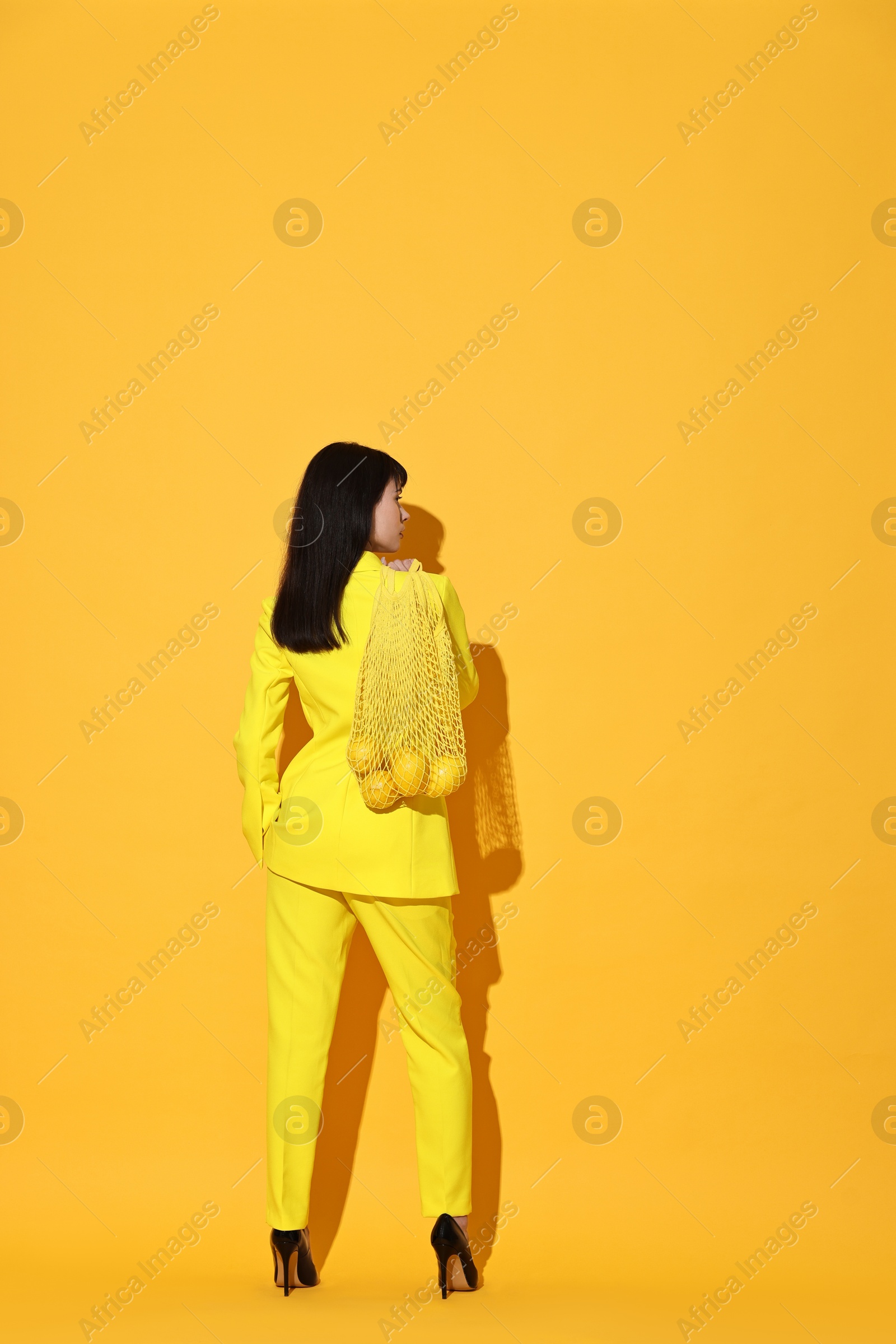 Photo of Woman with net bag and lemons on yellow background, back view