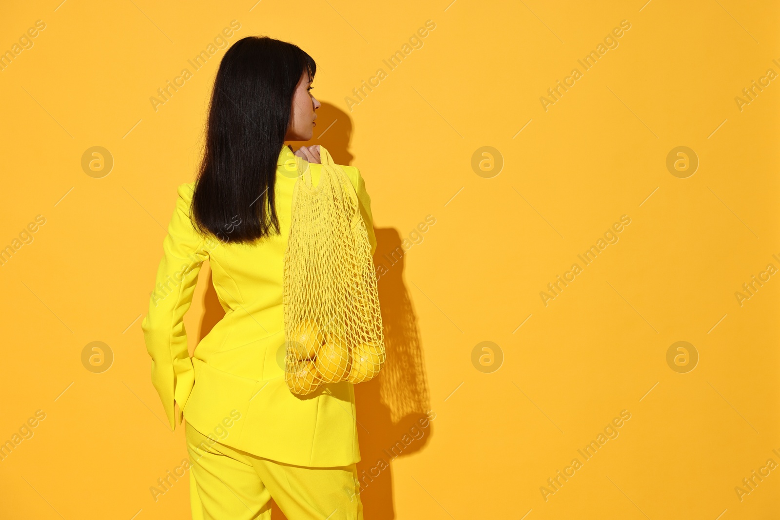 Photo of Woman with net bag and lemons on yellow background, back view. Space for text