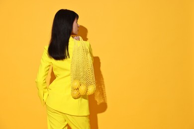 Photo of Woman with net bag and lemons on yellow background, back view. Space for text