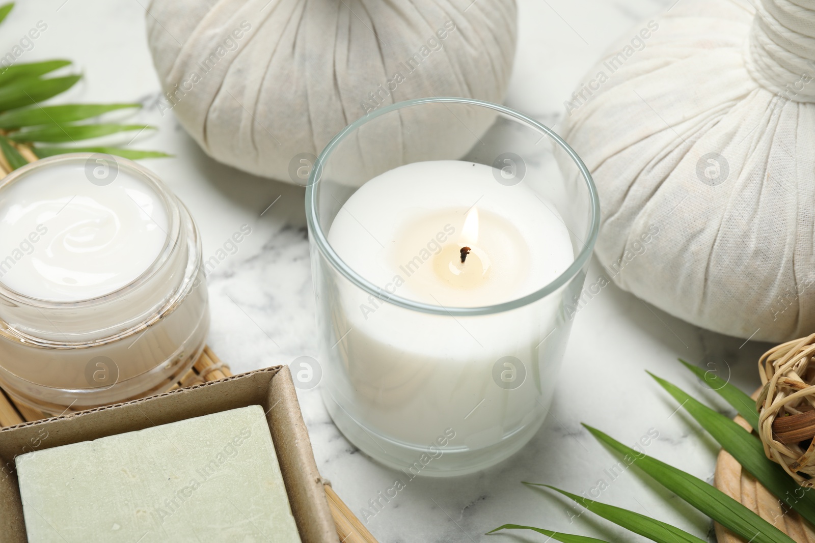 Photo of Spa composition with different cosmetic products, herbal bags, burning candle and leaves on white marble table, closeup