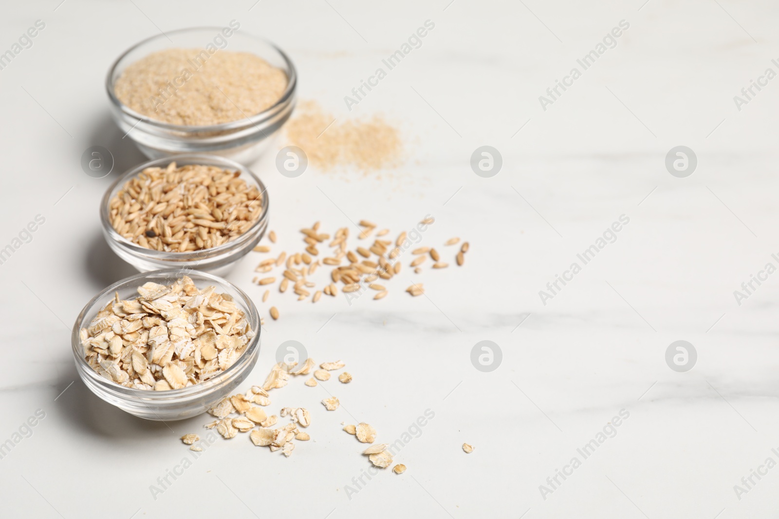 Photo of Oat bran, grains and flakes in bowls on white marble table, closeup. Space for text