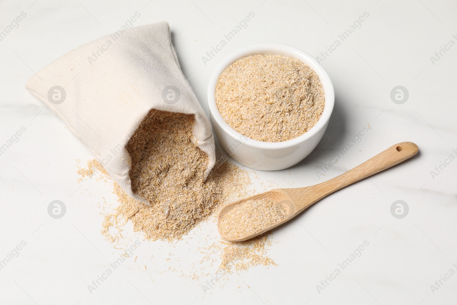 Photo of Oat bran in burlap bag, bowl and wooden spoon on white marble table