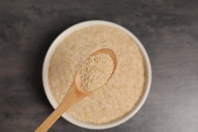 Oat bran in wooden spoon over bowl at grey table, top view