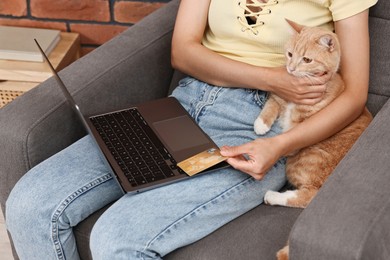 Online pet shop. Woman with laptop, credit card and cute cat making order on armchair at home, closeup