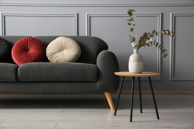 Photo of Stylish sofa with cushions and eucalyptus branches in vase on side table near grey wall