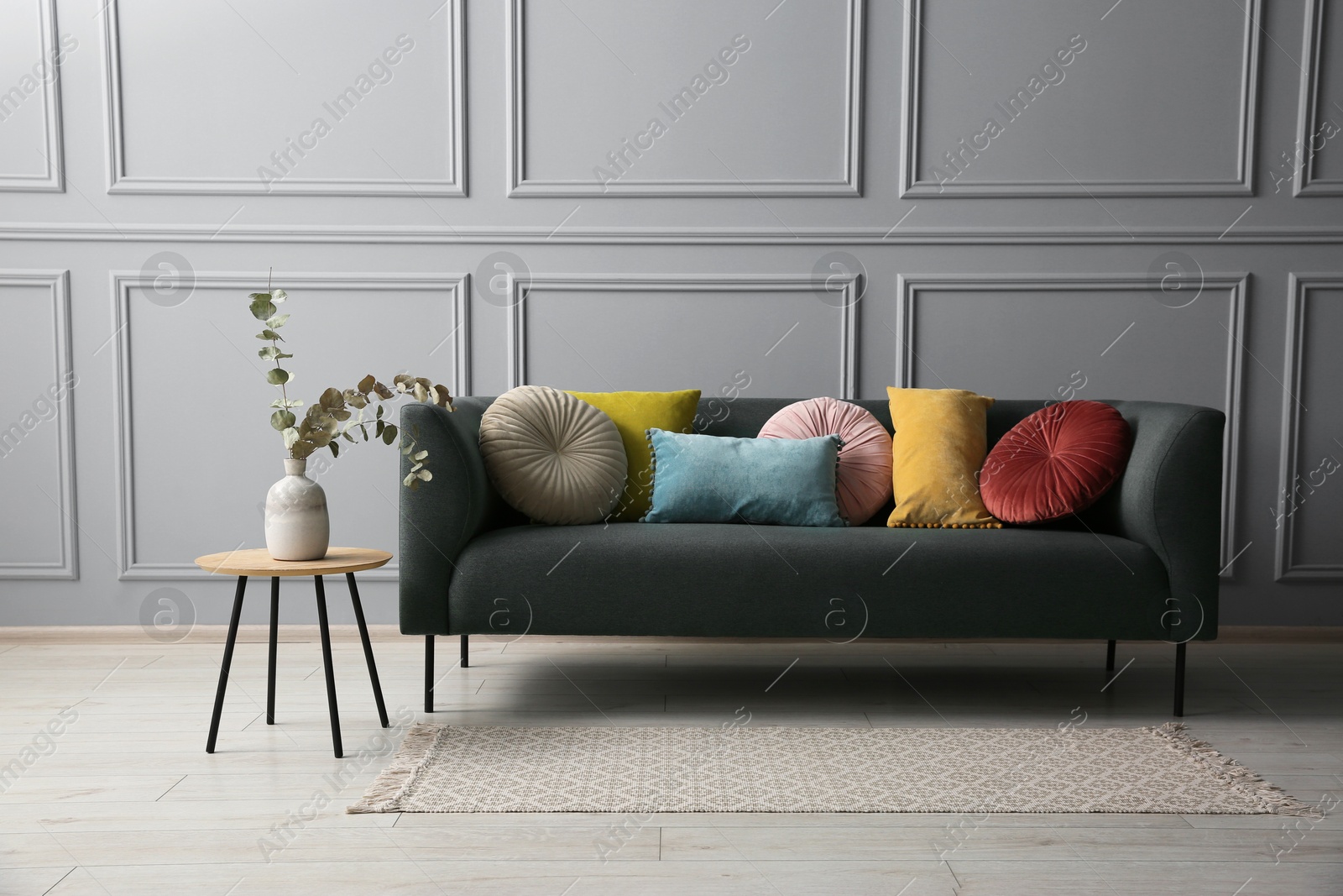 Photo of Stylish sofa with cushions and eucalyptus branches in vase on side table near grey wall