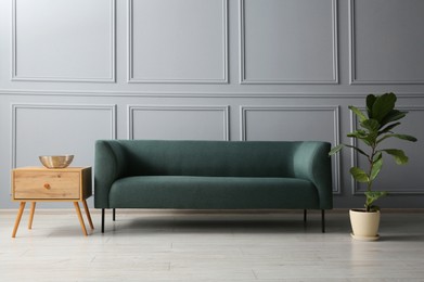 Photo of Stylish sofa, green plant and bowl on side table near grey wall