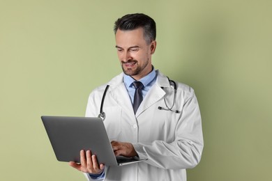 Smiling doctor with stethoscope using laptop on olive background