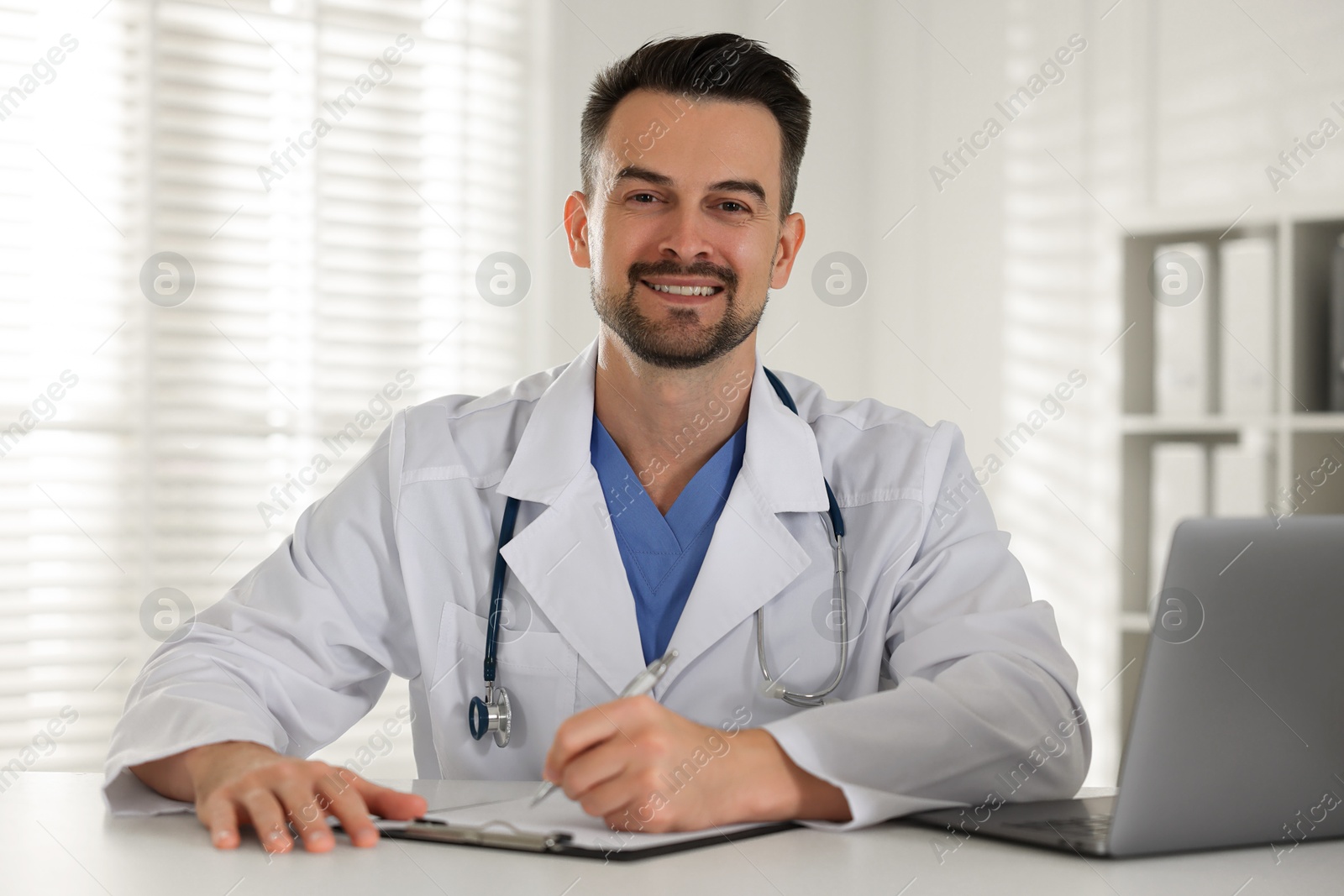 Photo of Smiling doctor with clipboard at table in clinic